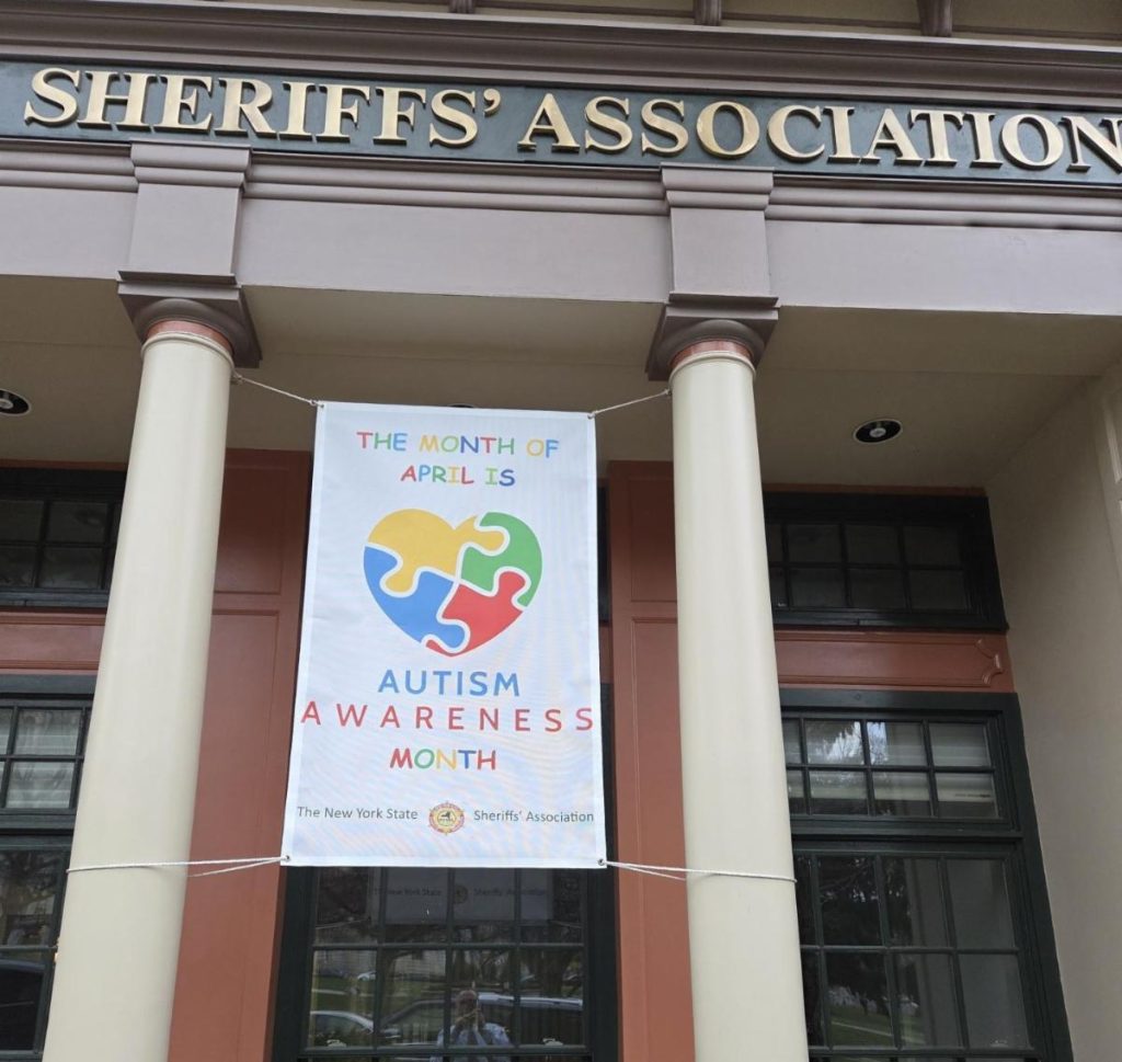 Photograph of a banner hanging between two columns outside a building labeled "Sheriffs' Association." The banner promotes April as Autism Awareness Month, featuring a heart-shaped puzzle piece design in blue, yellow, green, and red, with text in colorful letters and the New York State Sheriffs' Association logo at the bottom.
