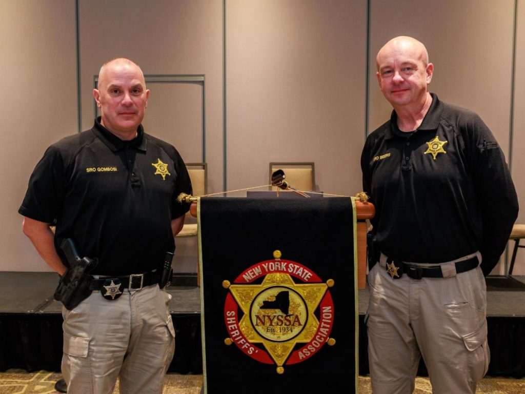 Photograph of two uniformed officers standing on either side of a podium draped with a New York State Sheriff's Association banner. The officers wear black shirts with gold star badges and beige pants, positioned in a conference or meeting room setting.