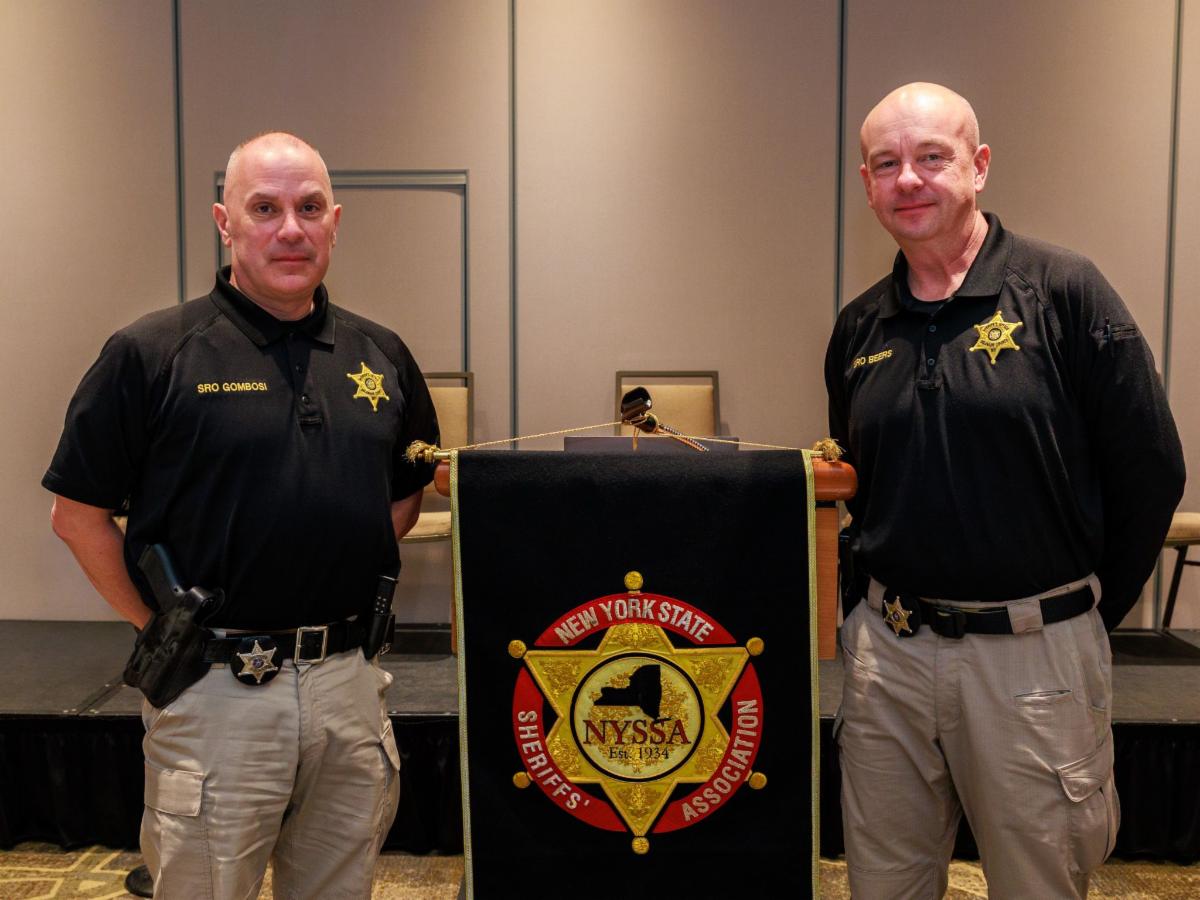 Photograph of two uniformed officers standing on either side of a podium draped with a New York State Sheriff's Association banner. The officers wear black shirts with gold star badges and beige pants, positioned in a conference or meeting room setting.