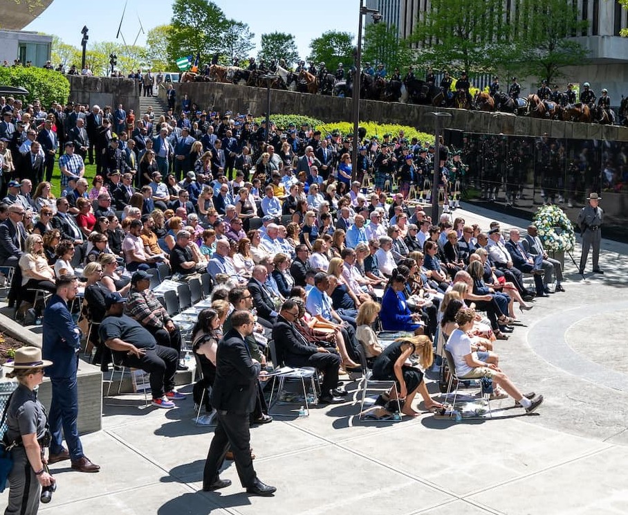 Photograph of a large outdoor gathering with people seated in rows of chairs and others standing behind them, attending a formal event or ceremony. The scene includes a speaker at a podium, uniformed officers, and a reflective black wall with a wreath, suggesting a memorial or commemorative occasion.