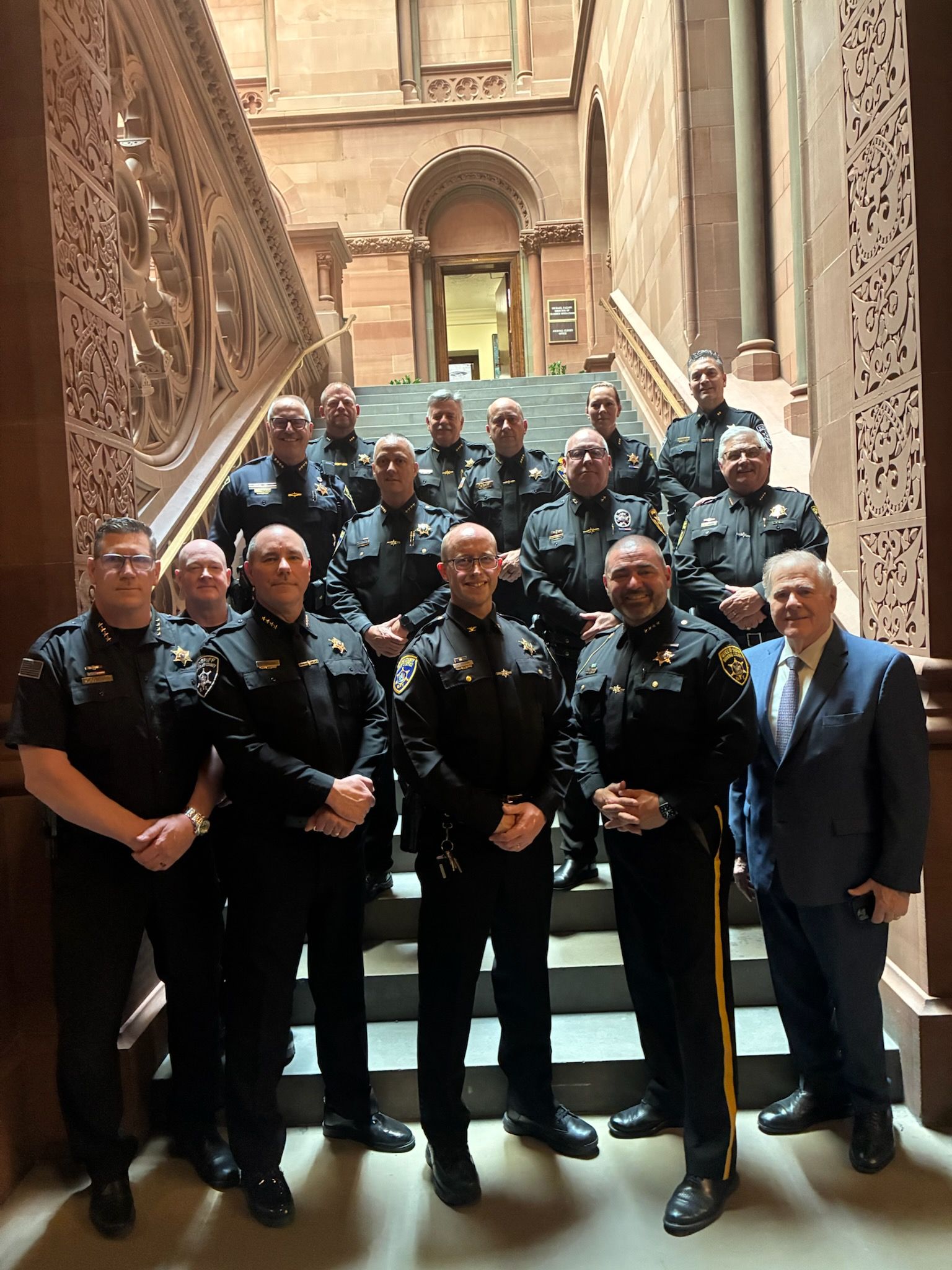 Photograph of a group of uniformed police officers and one person in a suit standing on a staircase inside a building with ornate stone walls and carved details. The group is arranged in rows, facing forward, with some officers wearing badges and patches visible on their uniforms.